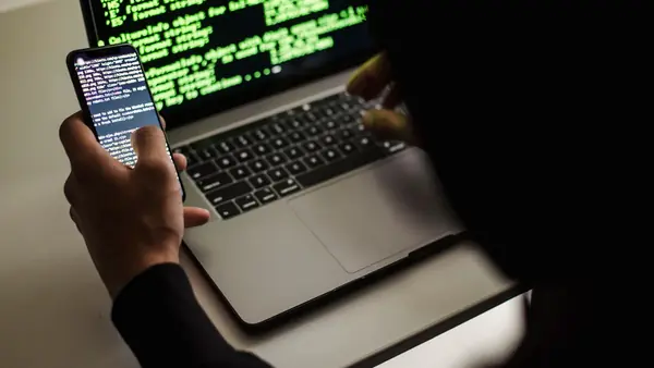 Person holding a smartphone in front of a laptop showing code in a dark room