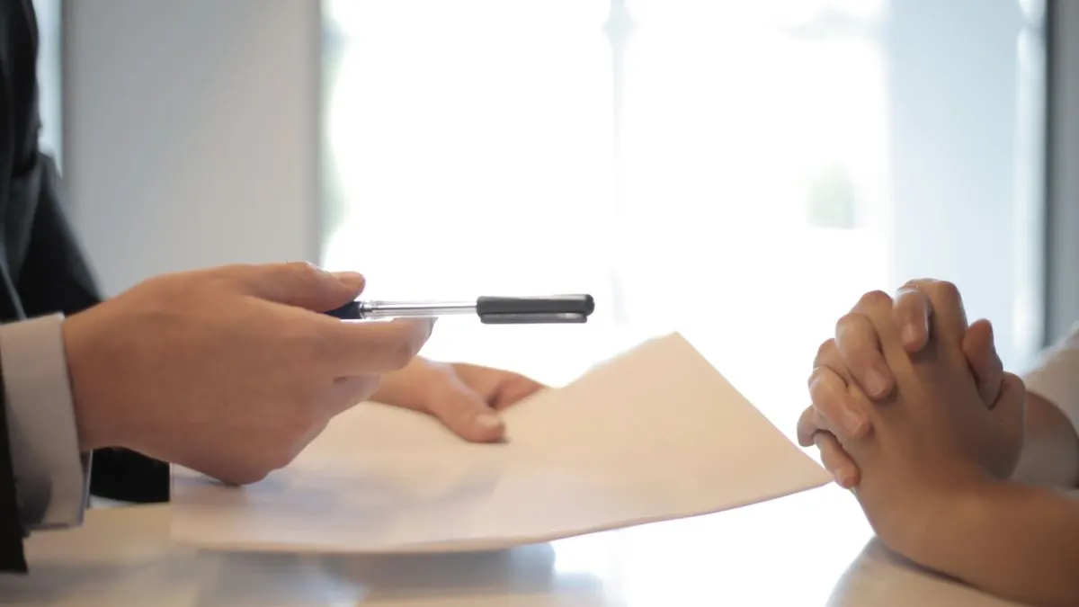 Hand passing a document and pen across a desk to a candidate